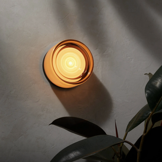 A sleek modern LED wall lamp with a round metal base, emitting a warm white glow, mounted on a wall beside some green foliage.