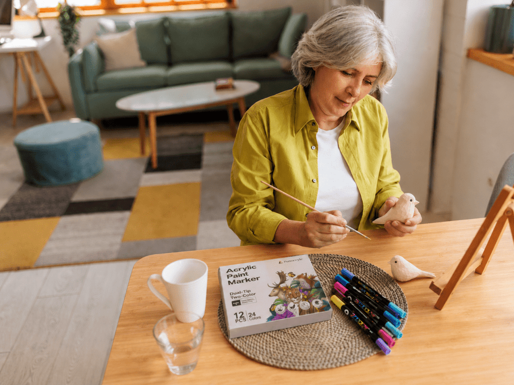 Older woman painting a wooden bird figurine at a table with acrylic paint markers