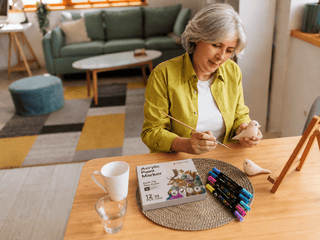 Older woman painting a wooden bird figurine at a table with acrylic paint markers