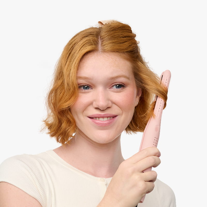 Woman with red hair using a pink hair straightener on a white background