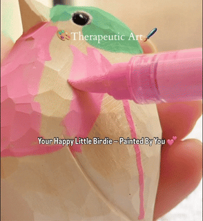 A person's hands using blue and pink acrylic paint markers to color a natural unfinished wooden bird, part of a DIY wooden animal coloring kit.